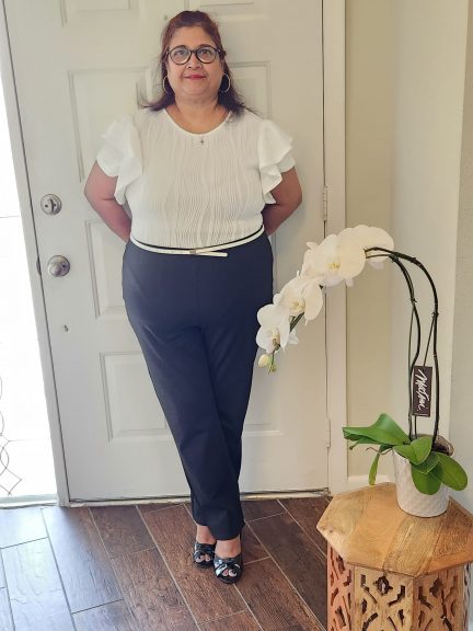 Plus-size woman in black pants and white bodysuit posing confidently indoors with black open-toe heels, standing beside a white orchid on a carved wooden table.