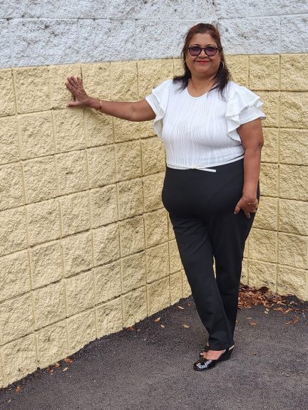 Plus size woman in a white ruffled bodysuit and black pants, posing against a two-toned yellow and white brick wall.