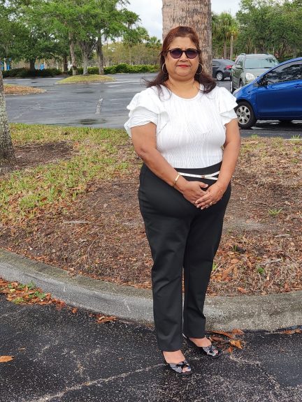 Plus-size woman in a white body suit and black pants standing outdoors in a parking area with trees and cars in the background.