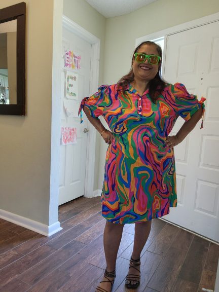 Plus-size woman standing confidently indoors wearing a short, multi-color abstract print dress with puff sleeves tied at the ends. She is smiling, hands on hips, and wearing bright multi-color sunglasses. The outfit is inspired by 1970's fashion. The background is a home hallway with warm lighting and wooden flooring.