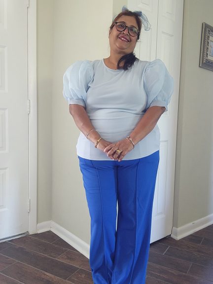 "A plus-size woman with dark hair and glasses stands frontally, wearing a light blue top with voluminous puff sleeves and a wide-leg blue pants. She has her hands clasped in front of her. The background shows a light-colored wall and two white doors."