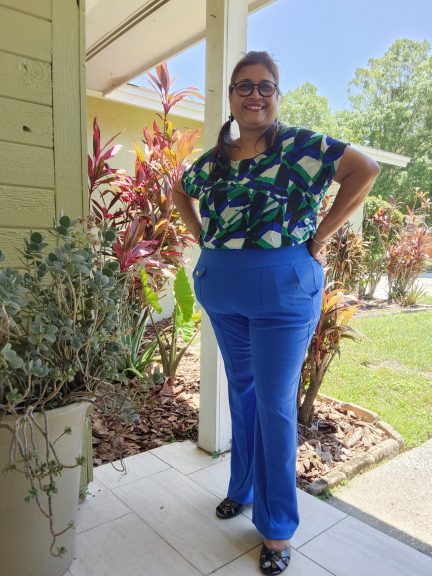 Plus-size woman standing confidently on a porch in daylight, wearing vibrant blue bell-bottom pants and a geometric print top in shades of green, white and navy. Her hair is styled in playful pigtails, and she is smiling brightly. The background includes plants, a garden bed, and trees under a clear blue sky.