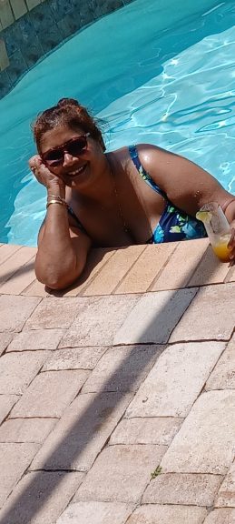 "A happy, confident plus-size woman in a blue floral swimsuit smiles while relaxing at the edge of a bright blue swimming pool, holding a refreshing drink."
