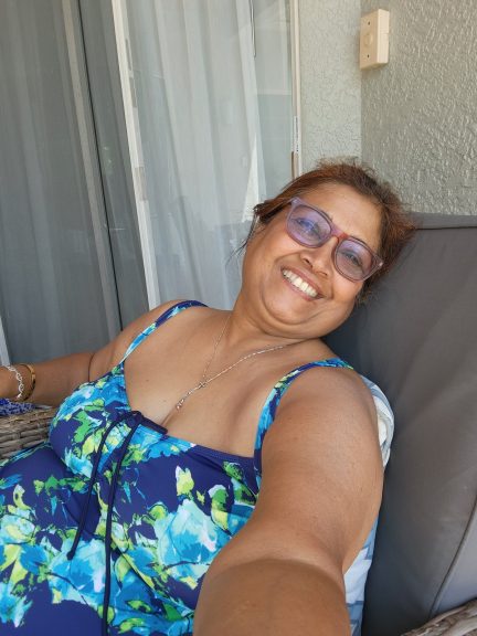 "A joyful plus-size woman in a blue floral swimsuit smiles broadly, relaxing comfortably in an outdoor chair on a balcony, embodying peace and self-care."